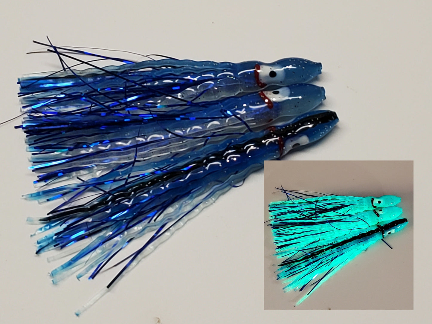 3 Blue with black back Glow Hoochie Squids with dark blue holographic Mylar Inserts laying on white background inset glowing in dark