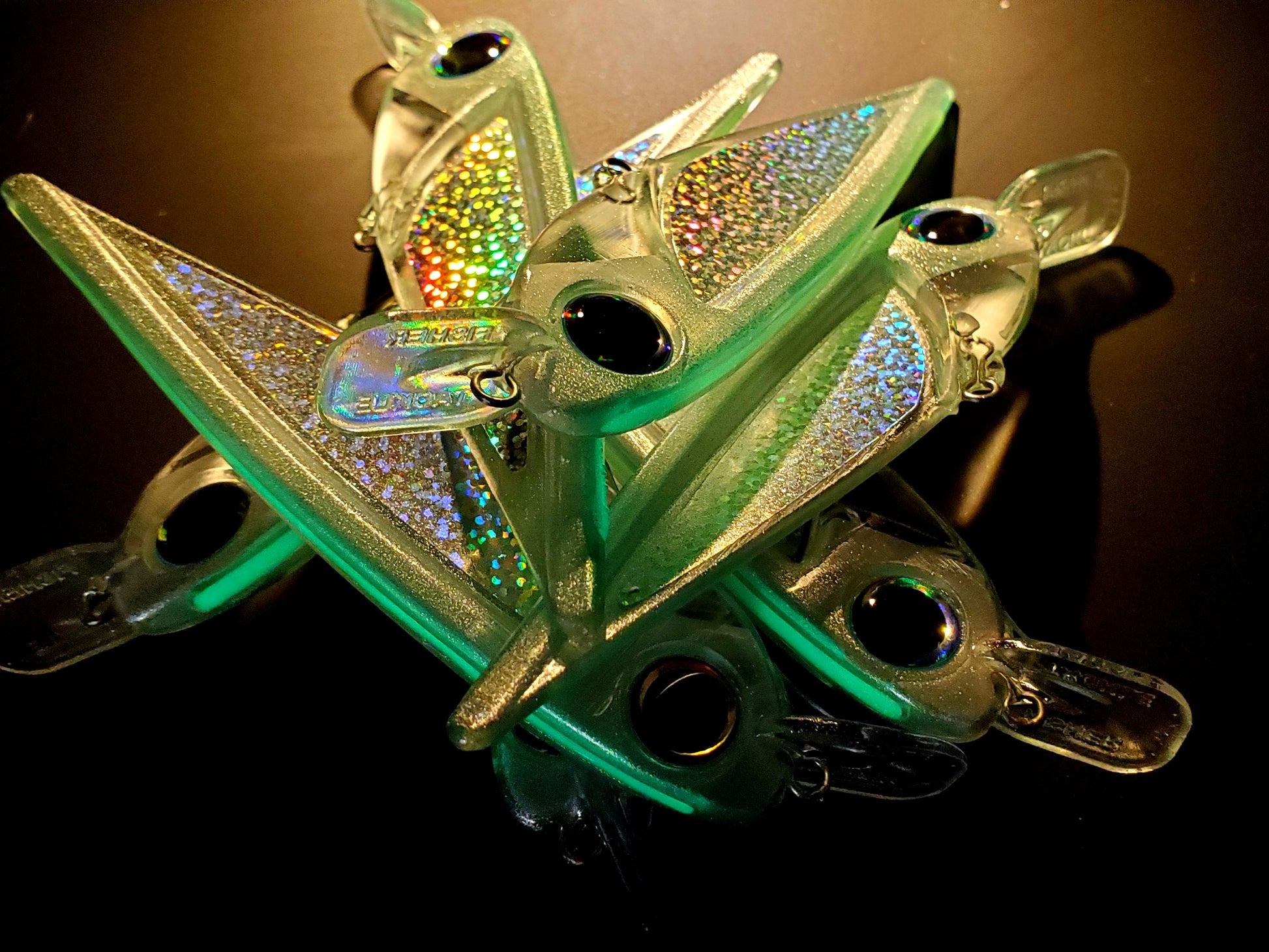 8pcs of sparkly greenish FlatFin FLASHBANG trolling lures in a pile on a black background with light from the back so some are glowing
