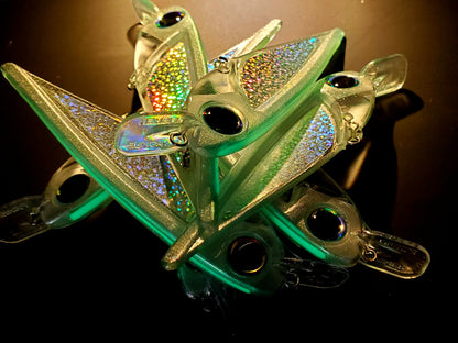 8pcs of sparkly greenish FlatFin FLASHBANG trolling lures in a pile on a black background with light from the back so some are glowing