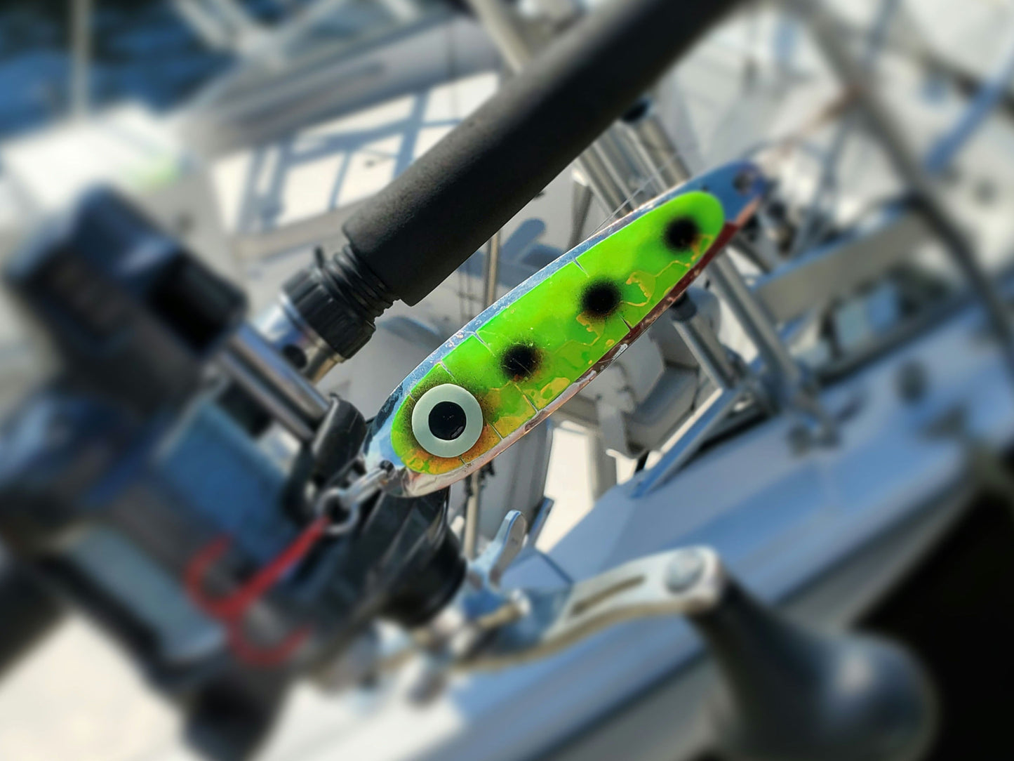 Close-up of a green with black dots fishing lure attached to fishing rod with a boat in background.