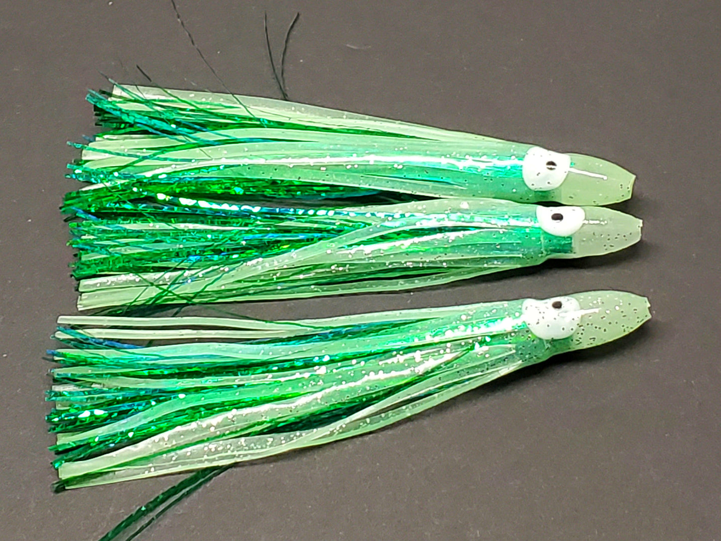 3 Pale green Glow Hoochie Squids with dark green holographic Mylar Inserts laying on dark background