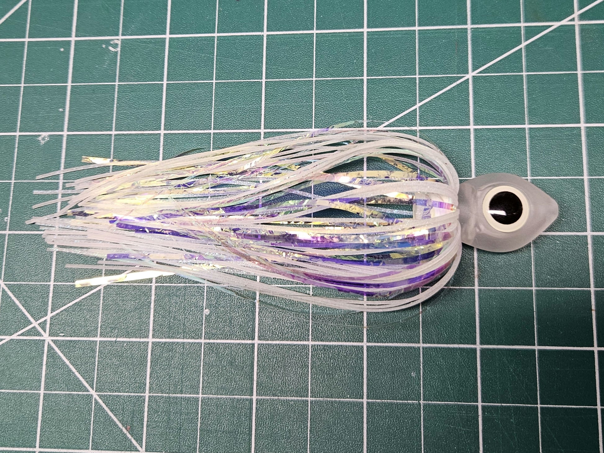 Side view of a Floating Squid Fly with a milky translucent hollow head, large black and glow eyes, and iridescent white and glow silicone skirting on a green mat.